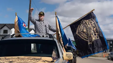 A separatist supporter waves a flag during as part of a convoy in Edmonton, Canada. The flag is black and says THE NORTH STANDS FREE above Alberta's crest, which contains mountains at the top and wheat at the bottom.
He is holding his fist up and is standing in the trunk of a moving pick-up truck. He is wearing a plaid shirt and a cap, with sunglasses.