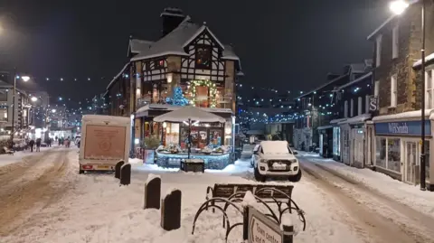 Snow covered town with lights strung up on a high street. 