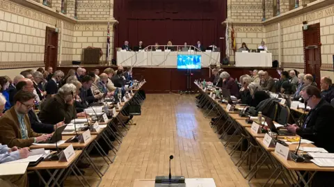 Lots of people sat at rows of individual brown tables, many wearing suits. Some people are sat at a table with a white tablecloth at the front of the room.