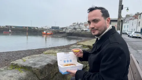 James Muirhead, who is wearing a dark coat, sits by a harbour on a grey day, holding a takeaway box of fish and chips and a lemon wedge. Fishing boats rest in the background near a stone pier, with pastel-coloured houses lining the waterfront.