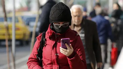 Anadolu via Getty Images A person in Iran holding a smartphone. They are wearing a big red jacket, a black hat, a black face mask and glasses.