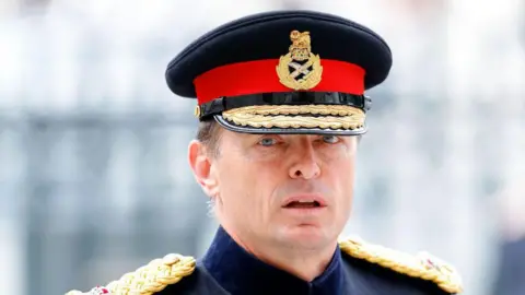 Getty Images A man in military regalia looks directly at the camera. He is wearing a captain's hat, with a red band and gold insignia on it. 