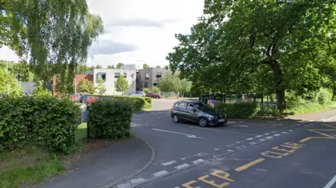 Google A driveway off a road leads towards a number of modern buildings in the distance. The road is lined with trees and hedges.