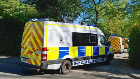Cheshire Police A police van enters a driveway situated between two hedges. It is following two other police vehicles.