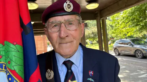 A man wearing a burgundy beret with glasses in a navy suit, looking into the camera. He is holding a blue, red and green regimental flag.