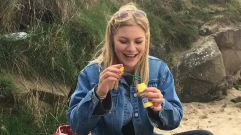 Ella Dawson Foundation A smiling young woman with long blonde hair and wearing a blue denim jacket sits on a sandy beach. She holds bubble mixture in a yellow container in her left hand, while preparing to dip the bubble wand into the solution with her right hand.