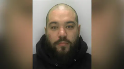 .Custody photograph of a person with a shaved head and beard wearing a dark hooded top against a plain background