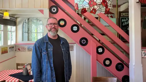 Owen Sennitt/BBC A man wearing a denim jacket with a beard and glasses stands inside an American-themed restaurant with a pink staircase and neon signs