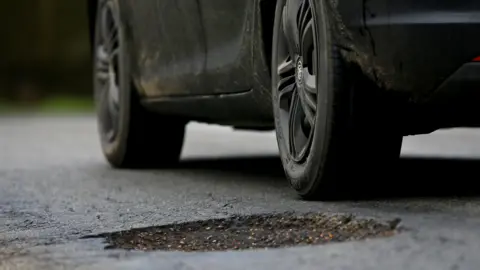 A black car parked with its rear wheel next to a large pothole in the road surface.