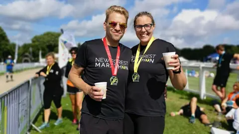 The image shows a picture of a man and woman posing for a photograph, both holding a white cup each and with a medal around their necks. The man is wearing sunglasses while the woman is wearing regular glasses. People can be seen in the background standing and lying down on grass.