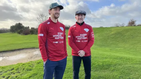 Tom Lockyer and Ian Holloway stand on a golf course and wear red sport jackets advertising the every minute matters campaign. Holloway stands on the right and wears a flat cap and looks at the camera. Lockyer is on the left with his hands in his pockets and wearing a green baseball cap.