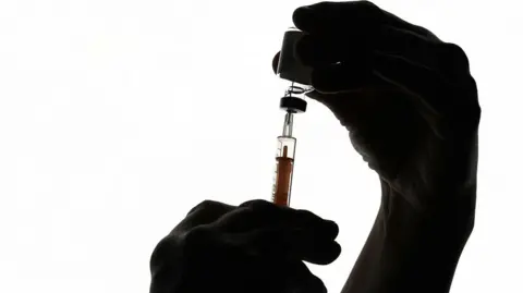 PA Media A picture of hands holding a needle and bottle. It is on a white background. 