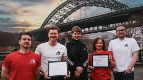 Thomas Jackson The Sunderland Music City Team – Marty Longstaff (left, in red Sunderland Music City T-shirt), Frankie Francis (centre left, in white Sunderland Music City T-shirt), Michelle Daurat (centre right, in red Sunderland Music City T-shirt) and Andrew Dipper (right, in white Sunderland Music City T-shirt) with managing director of the Music Cities Network, Lena Ingwersen (centre, in black Adidas tracksuit with three white stripes across the sleeves) and holding the membership certificate. Marty and Frankie have short brown hair, swept to the back. Michelle has shoulder-length straight brown hair with a side fringe. Andrew is wearing glasses and has a buzz cut. Lena has short wavy brown hair with a fringe. Sunderland's Wearmouth Bridge is in the background. It is a green arch structure.