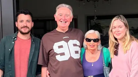 পল ওটেসেন চারজন একে অপরের পাশে দাঁড়িয়ে আছে। বাম থেকে ডানে – দাড়ি-গোঁফ ও কালো চুলওয়ালা একজন লোক লাল টি-শার্ট এবং সবুজ শার্ট পরা। তার পাশে ধূসর চুলের একজন বৃদ্ধ লোক, একটি বাদামী টি-শার্ট পরা যার গায়ে 96 নম্বর সাদা লেখা। তার পাশে লম্বা সাদা চুলের একজন বয়স্ক মহিলা, সানগ্লাস পরা। তিনি একটি বেল্ট এবং একটি সবুজ কার্ডিগান সহ একটি বেগুনি টপ পরেছেন। শেষে লম্বা স্বর্ণকেশী চুল এবং কলার থেকে ঝুলন্ত সানগ্লাস সহ একটি গোলাপী টপ সহ এক যুবতী।