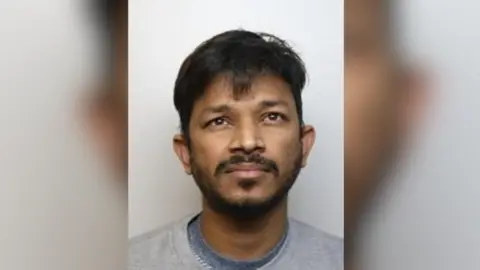 A custody image of Trevor Fernandes. He is wearing a grey prison tracksuit and looking to the top left of the screen.