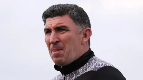Kevin Maher has short, dark hair. He is licking his lips with a serious expression on his face. He is wearing a black club tracksuit with white detailing on the shoulders.