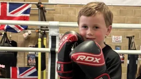 Supplied Jesse standing inside a boxing ring at a gym. He is wearing red and black gloves and holding them up in a fighting stance. He is smiling.