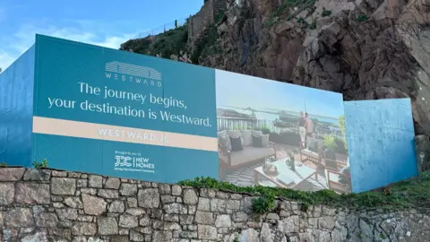 A picture of a sign with a brick wall below it. The sign has an image of a property on it, next to writing. It is blue in colour.