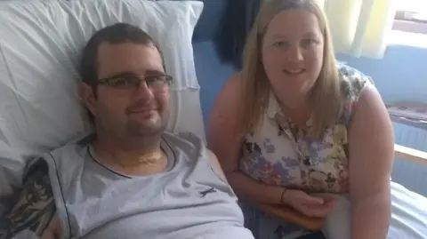 Steve Jones Steve Jones sitting up in a hospital bed wearing a grey sleeveless top and glasses. On the right, leaning over a chair arm, is Claire Jones who has straight fair hair over her shoulders and is earing a floral top. Ward J2 at Addenbrooke's Hospital, August 2018