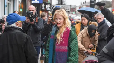 PA Media MP Hannah Spencer, wearing a green coat and multicoloured scarf is stood on a pavement surrounded by photographers as she gives an interview to a man in a blue baseball cap and black jacket.