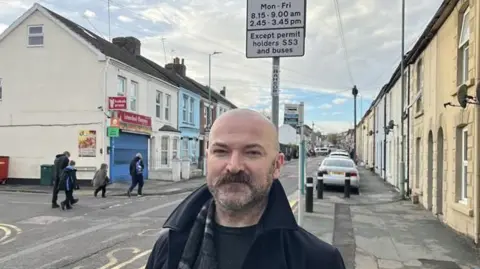 Councillor Alex Paterson from Medway Council standing on Richmond Road, Gillingham next to a School Streets Scheme restrictions sign as children walk to school on the opposite footpath