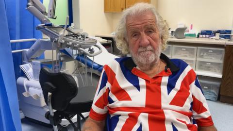 Geoff Le Gallez with grey hair and beard, sat in a clinic wearing a shirt with a pattern of the Union Flag