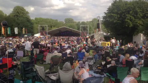 A large crowd, many sitting in camping chairs face towards a stage at a festival. 