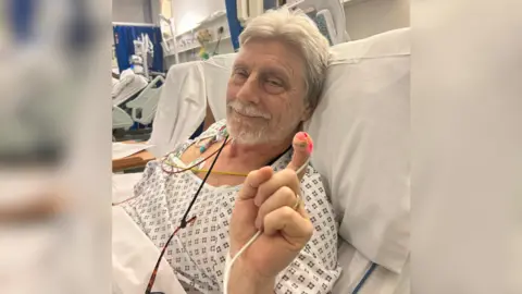 The image shows an elderly man lying in a hospital bed wearing a hospital gown' there are tubes coming out of his shoulder and one connected to his finger, which he is holding up.