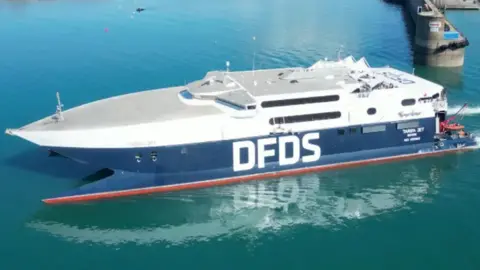 The picture shows a large high-speed ferry on calm blue water near a dock. The vessel has a sleek, modern design with a pointed bow and a flat upper deck. It is painted mainly white on the upper section and dark blue on the lower section, with the letters DFDS prominently displayed in bold white font along the side.