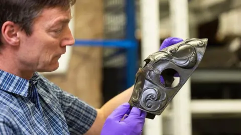 A man in a checked shirt wearing purple gloves carefully holds a Celtic ceremonial artwork in his hands