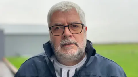 A man with a beard, glasses and white hair is looking into the camera. He is wearing a blue rain jacket. 