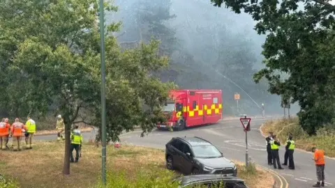 Emergency service workers in hi-vis jackets congregate in two groups - one group of police and one group of firefighters, in the road next to a fire engine. There are two parked cars between the group. Thick smoke is visible between the trees.