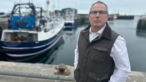 Mike Park looking straight at the camera. He is wearing a dark green gilet with a brown collar over a white shirt. He is wearing glasses and is standing in front of a harbour with boats in it.