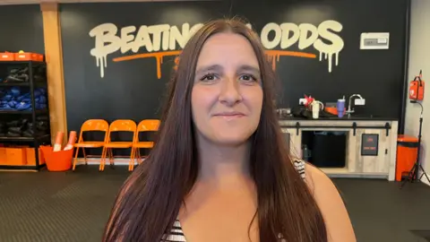 Lisa has long dark hair and is standing in the gym. A black wall behind has white writing which read 'beating odds"