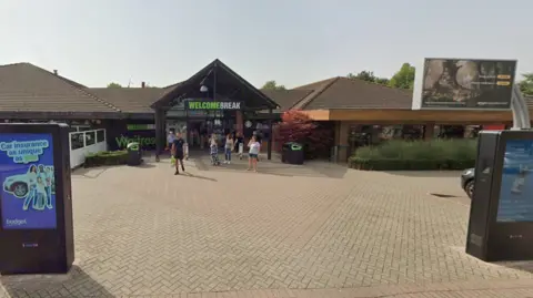 Google Maps A Welcome Break service station on a sunny day. The service station is a one-storey building with a triangular roof, and there is a sign saying "WELCOME BREAK" in green and white capital letters on the front of the building.