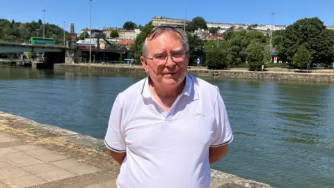 Mike Ferguson standing by the River Avon along Bristol's Harbourside. He is wearing a white polo-shirt and has short grey hair and black glasses.