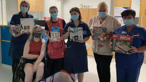 Katie Callaghan A group of nurses are holding books and art supplies in a hospital. Katie is sat nearby. Everybody is wearing a face mask.