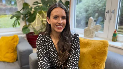 Rebecca Thomas, a teacher, sitting on a sofa. She is wearing a black and white top.