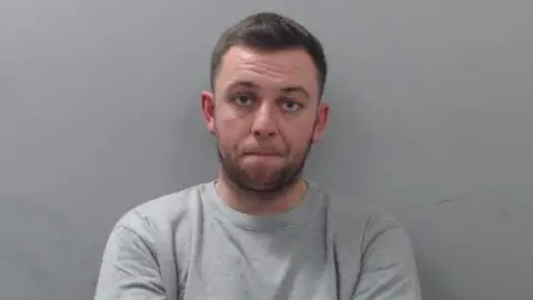An image of Matthew Brogan sitting in front of a grey wall. He has short brown hair and a beard. He is looking at the camera with a neutral expression on his face. He is wearing a light grey, crewneck jumper.