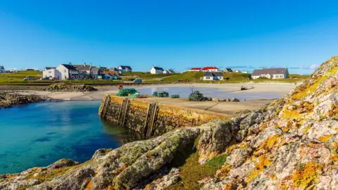 White-walled properties near a harbour. There are lichen-covered rocks in the foreground and the picture 