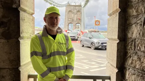 A middle-aged man wearing yellow hi-vis on a large bridge crossing. There's cars travelling in both directions.