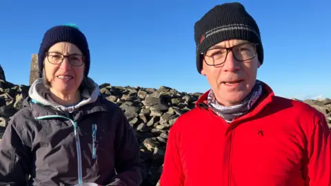 Mary and James outside wearing hiking gear and hats. Both are wearing black framed glasses. 