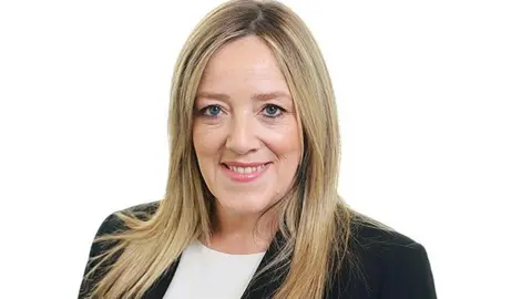 A smiling woman with log blonde hair and wearing a navy jacket.