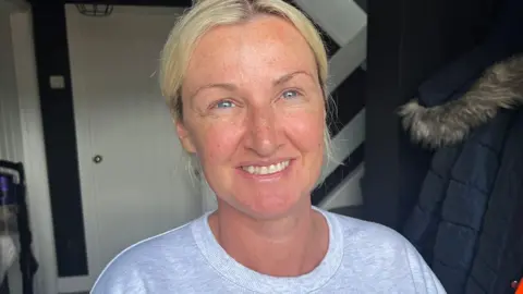Head and shoulders of Kerry Robinson standing in the doorway of her home. She has blonde hair, parted down the centre and tied back and is wearing a pale grey tshirt and smiling