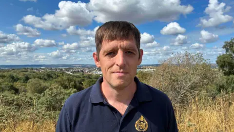 A close-up of a man with brown hair looking at the camera with a solemn expression, wearing a blue polo shirt that says FBU on it in gold thread. He stands in front of shrubbery and trees.