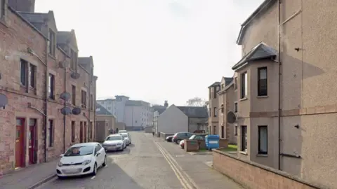 Google A residential street with flats on either side and cars parked along the road. A hotel building is at the end of the road in the background of the shot.
