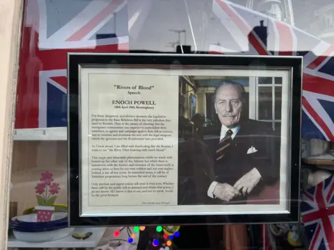 A framed image of a man in a suit and tie next to a text of Enoch Powell's "Rivers of Blood" speech. There are Union flags in the background.