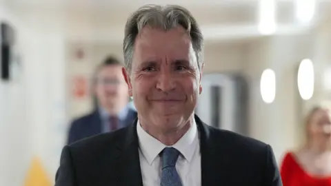 A close-up picture of Dan Norris, who is walking down a bright corridor. He has short grey hair and is smiling slightly. He is wearing a dark suit with a blue tie and white shirt. 