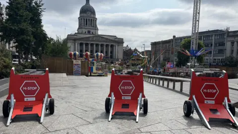 Three red barriers with H-Stop written on them, lined up next to Nottingham's Old Market Square with the Council House in the background