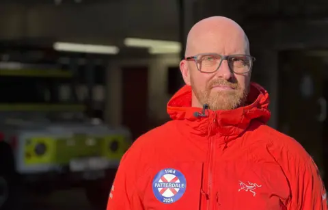 Matthew Cox is wearing a red Patterdale Mountain Rescue jacket and is standing in front of a mountain rescue vehicle. 